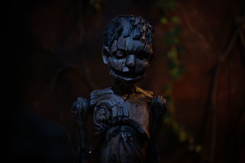 La espeluznante marioneta de madera de Pinocho tallada rústicamente, parada en la penumbra con ojos fijos y sonrisa siniestra.