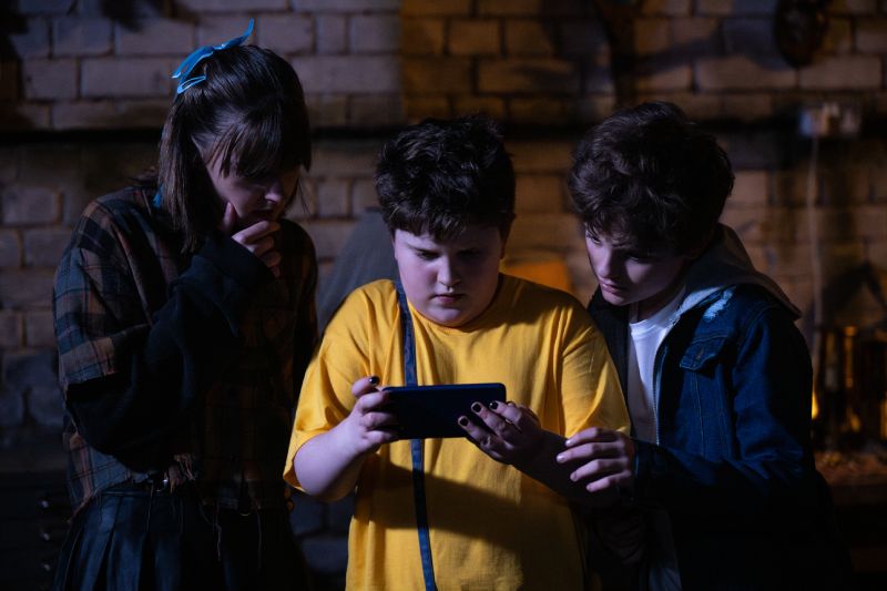 Tres niños reunidos en un sótano iluminados por la luz de una pantalla de teléfono celular que sostienen en el centro.