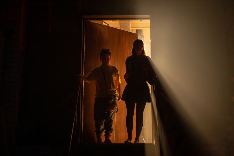 Siluetas de dos jóvenes entrando por una puerta iluminada por un fuerte haz de luz contraluz desde el exterior.