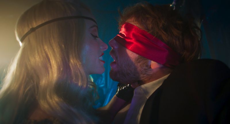 Una mujer rubia con venda roja en los ojos e inclinada hacia adelante compartiendo un momento de tensión y cercanía con un hombre.