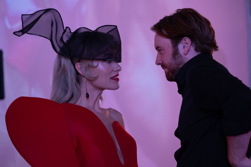 Olivia Wilde luciendo un vestido rojo con un tocado negro de malla junto a Cooper Hoffman en una habitación iluminada en tonos rosados.