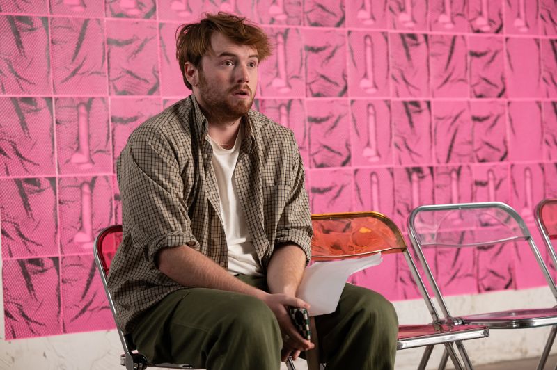 Cooper Hoffman sentado con expresión de desconcierto frente a una pared decorada con patrones rosados.