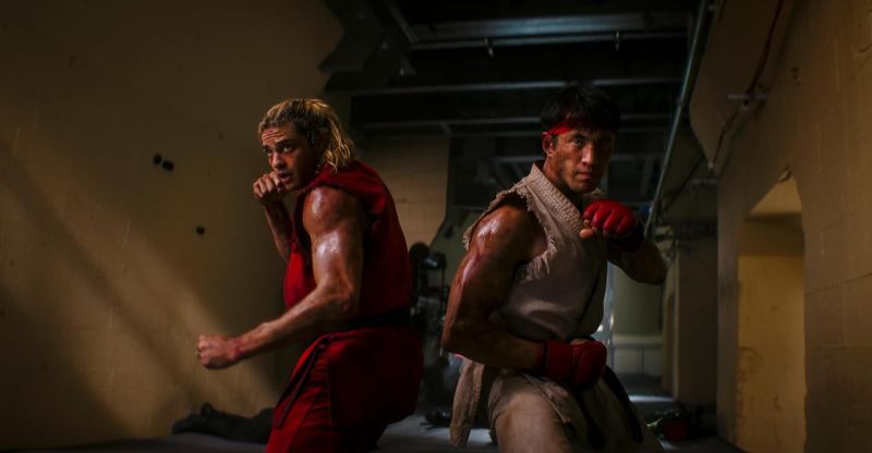 Andrew Koji como Ryu y Noah Centineo como Ken Masters colocados espalda con espalda en postura de combate dentro de un pasillo industrial iluminado de forma tenue.