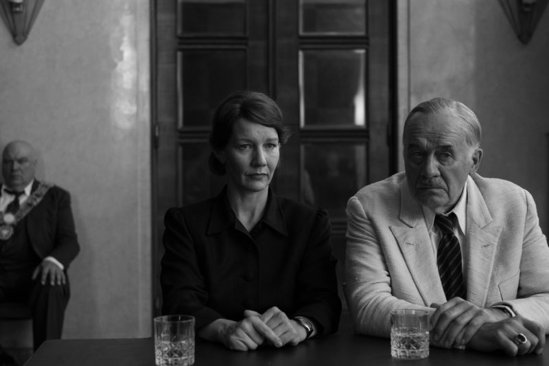 Tensiones Familiares e Intelectuales en la Trama Hanns Zischler como Thomas Mann y Sandra Hüller sentados con expresión seria frente a una mesa con vasos de cristal en una sala de reuniones.
