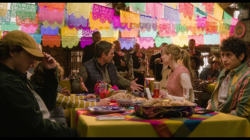 Escena de discusión en una mesa de restaurante decorada con papel picado y un mariachi tocando al fondo.