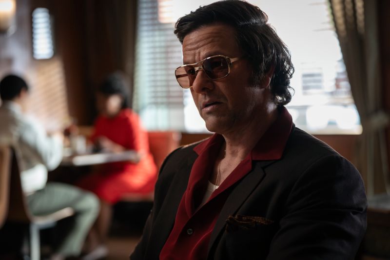 Plano medio de Mark Wahlberg interpretando a Greg Scarpa con lentes de sol de armazón dorado, peinado retro y camisa roja desabotonada bajo un saco.