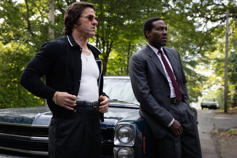 Mark Wahlberg y Yahya Abdul-Mateen II recargados al frente de un automóvil clásico azul en un camino rodeado de árboles.