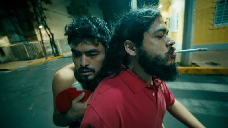 Recorrido nocturno en motocicleta en A toda madre Dos hombres en una motocicleta en movimiento de noche por la calle; el conductor tiene barba y un cigarrillo encendido en la boca mientras el pasajero lo abraza tenso por la espalda.