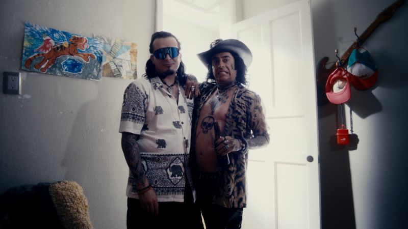 Personajes extravagantes en una escena de A toda madre Dos hombres con vestimenta estrafalaria, tatuajes visibles, sombreros y lentes oscuros posando juntos en el marco de la puerta de una habitación.