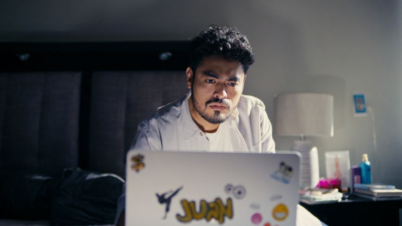 Momento de revelación frente a la computadora en A toda madre Un hombre sentado en la cama frente a una computadora portátil con calcomanías, mirando fijamente la pantalla con expresión de preocupación y concentración en una habitación penumbrosa.