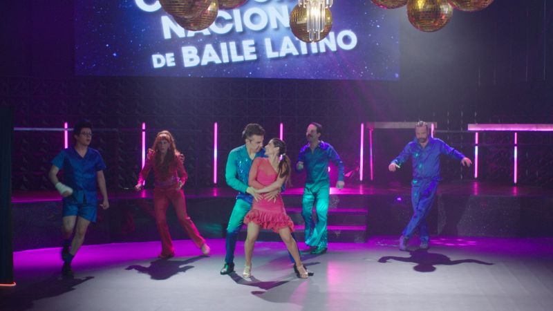 Presentación del grupo en la Competencia Nacional de Baile Latino Los miembros del club bailando en pareja vestidos con trajes brillantes y vistosos sobre el escenario iluminado de la Competencia Nacional de Baile Latino.