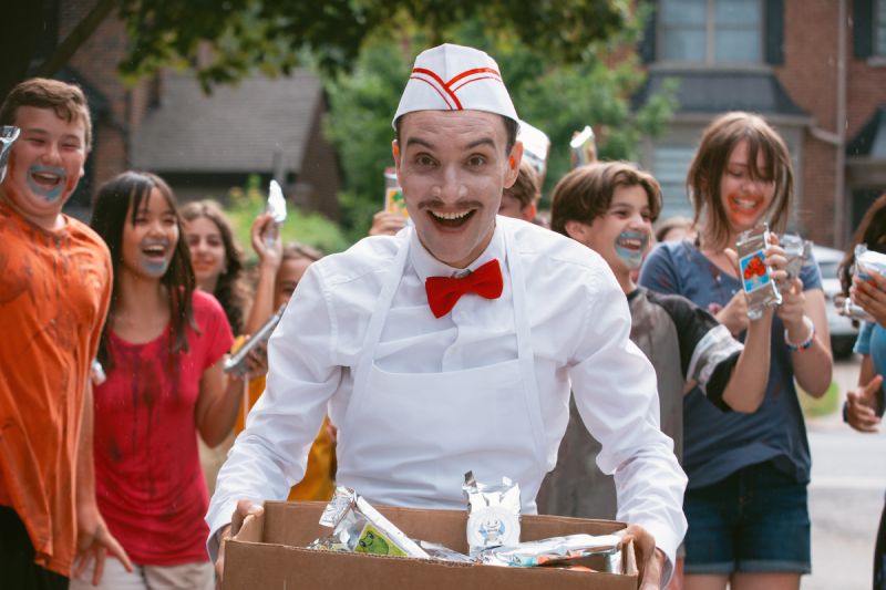 El siniestro heladero repartiendo paletas y dulces El actor caracterizado como el alegre y siniestro heladero sonríe eufóricamente mientras viste un uniforme blanco con moño rojo, cargando una caja de golosinas rodeado de niños entusiasmados.