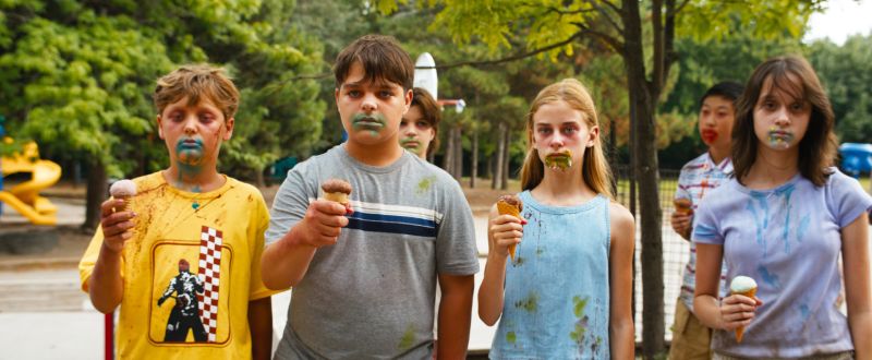 Niños bajo el influjo del helado en un parque Cuatro niños parados en línea en un parque sosteniendo conos de helado, con miradas perdidas y expresiones catatónicas, con manchones coloridos y oscuros alrededor de sus bocas.