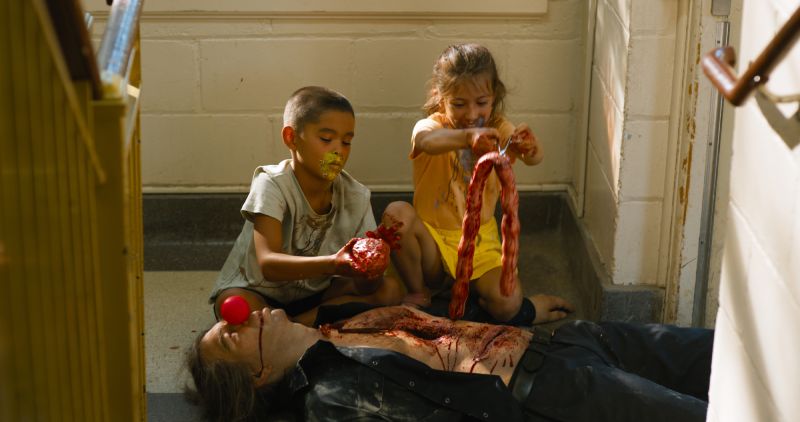 Escena de violencia explícita con niños y vísceras Dos niños pequeños sentados en el suelo jugando violentamente con las vísceras e intestinos de un hombre sin vida tirado a su lado con una nariz de payaso.