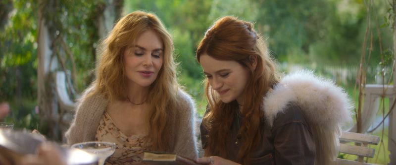 Nicole Kidman en suéter beige junto a Joey King leyendo sonrientes un libro antiguo sobre una mesa en el jardín rodeadas de vegetación.