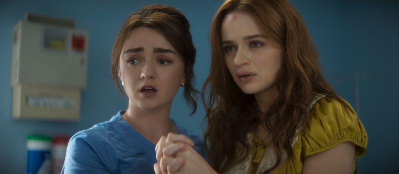 Maisie Williams vestida con uniforme médico azul sosteniendo con preocupación la mano de Joey King en una habitación de hospital.