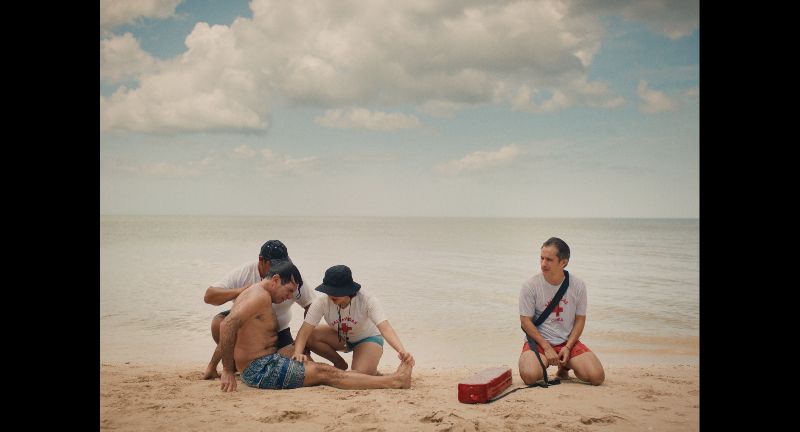 Escena del rescate en la playa en Hombre al agua Fotograma de la película mostrando a un hombre rescatado en la playa siendo atendido por paramédicos y un salvavidas sentado al lado sobre la arena.