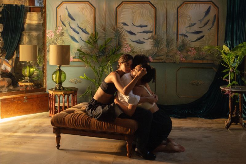 Momento de tensión familiar en Hombre al agua Fotograma dramático de tres personajes abrazados en el suelo de una habitación elegante con decoración tropical.