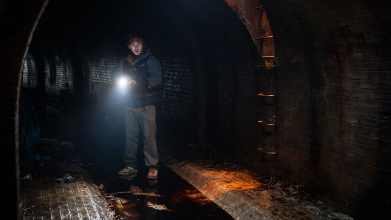 Bryan (Austin Abrams) parado en medio de un túnel o drenaje subterráneo oscuro de ladrillo, sosteniendo una linterna encendida que ilumina el ambiente húmedo y sombrío.