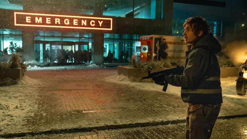 Bryan parado afuera de la entrada iluminada de emergencias de un hospital en medio de la nieve, sosteniendo un arma subametralladora mientras mira hacia los lados alerta.