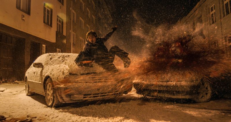 Escena de acción nocturna en la calle cubierta de nieve donde Bryan sale disparado e impacta sobre el cofre de un automóvil durante una violenta explosión o colisión.