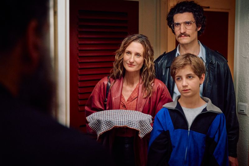 Vincent junto a sus padres en un momento de visita familiar Vincent (Simon Boublil) viste una chamarra deportiva azul junto a sus padres (Camille Cottin y Louis Garrel) frente a una puerta de madera sosteniendo una bandeja cubierta.