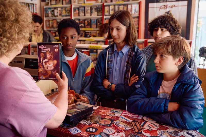 Vincent y sus amigos visitando el videoclub de la colonia Cuatro jóvenes adolescentes frente al mostrador de un videoclub ochentoso mirando con curiosidad una cinta de VHS que sostiene la encargada.