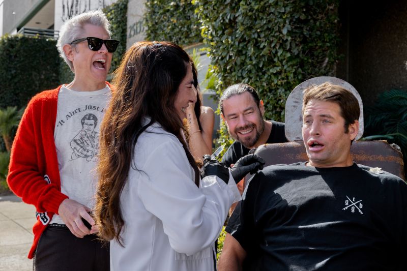 Fotografía promocional de Jackass: La Última y Nos Vamos donde Johnny Knoxville, vistiendo un suéter rojo y anteojos de sol, ríe efusivamente junto a Chris Pontius mientras una especialista médica aplica una inyección en el labio de un participante sentado en una silla.