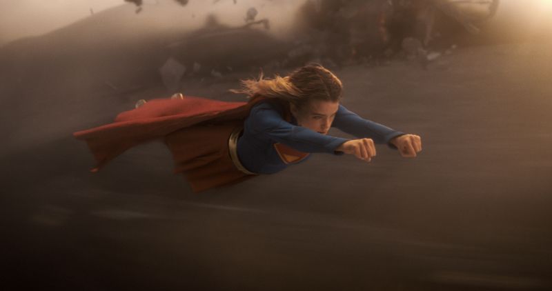 Secuencia de acción que muestra a Supergirl volando a gran velocidad a baja altura a través de un entorno desértico envuelto en polvo y viento.