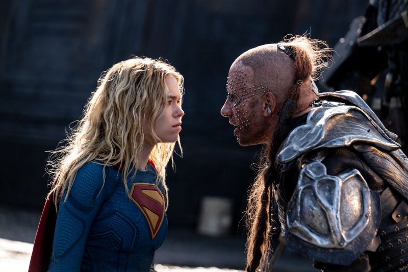 Plano medio de un tenso enfrentamiento cara a cara entre Supergirl (Milly Alcock) y un temible cazador de recompensas alienígena con implantes metálicos en el rostro.