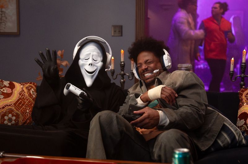 Un hombre sentado en un sillón junto a un personaje con la túnica negra y máscara sonriente de Ghostface. Ambos sostienen mandos de consola de videojuegos y llevan auriculares blancos con micrófono integrados, riendo en una habitación decorada con velas sobre candelabros.