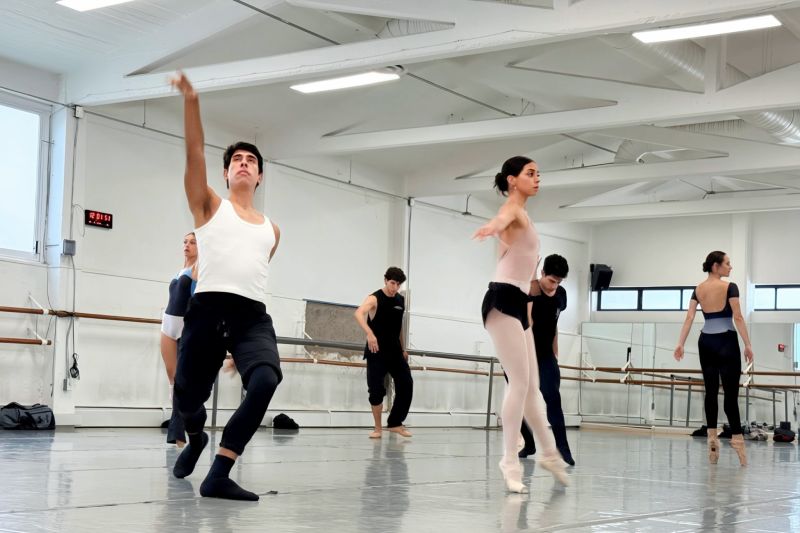 Fotografía de ensayo en salón de danza iluminado donde varios bailarines y bailarinas de la Compañía Nacional de Danza realizan movimientos contemporáneos y clásicos; en primer plano un bailarín con camiseta blanca extiende el brazo hacia arriba mientras una bailarina en puntas realiza una postura grácil.