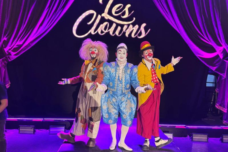 Les Clowns de la Carpa Astros y Circo Atayde llega a Texcoco Tres artistas de clown caracterizados con vestuarios coloridos y maquillaje posan sobre el escenario frente a un fondo negro con el nombre del espectáculo 'Les Clowns' iluminado al centro y cortinas púrpuras a los costados. El personaje de la izquierda viste traje parcheado de tonos marrón y peluca rubia despeinada; el del centro luce un elegante traje azul claro de época con bordados dorados y rostro maquillado de blanco; y el de la derecha viste saco amarillo, pantalón rojo y sombrero rojo.