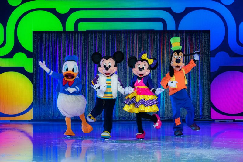 Mickey, Minnie, Donald y Goofy posan juntos en un escenario iluminado con patrones multicolores.