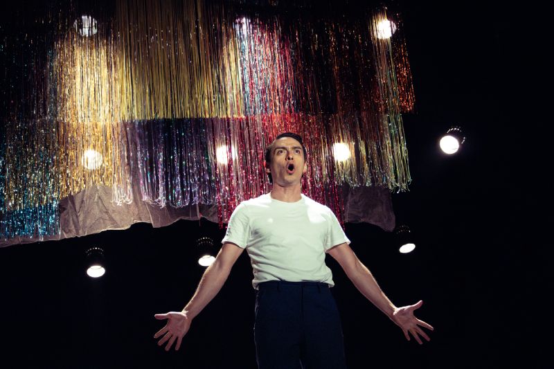 Persona sobre un escenario iluminado por reflectores, con brazos extendidos y decoraciones metálicas suspendidas en el fondo.