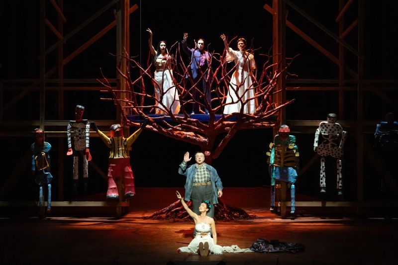 Escena teatral con un árbol escultórico rojo en el centro, rodeado de intérpretes y figuras humanas estilizadas en un escenario iluminado.