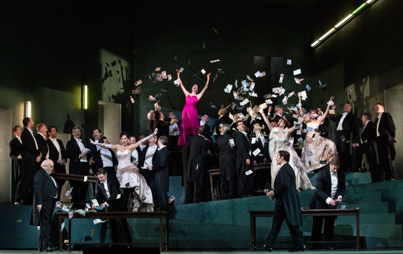 Fotografía de la puesta en escena de la ópera Manon sobre el escenario del Metropolitan Opera House. En el centro, una solista con vestido de gala rosa celebra mientras billetes de papel caen sobre el elenco distribuido en una escenografía inclinada de tonos verdes y elegantes trajes de etiqueta.