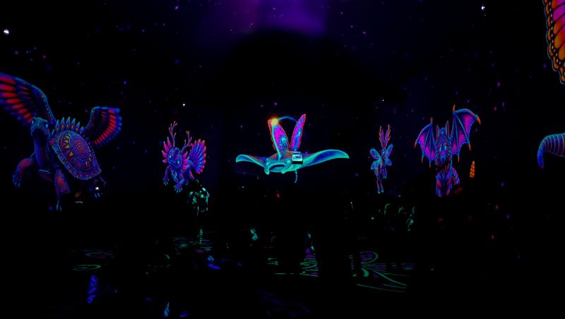 Proyección inmersiva en un espacio oscuro donde destacan alebrijes y figuras mitológicas voladoras iluminadas en tonos fluorescentes neón como morado, azul, rosa y naranja. Entre las criaturas se distinguen un ave con alas desplegadas, una mantarraya brillante y un mamífero alado rodeados de patrones prehispánicos en un entorno envolvente.