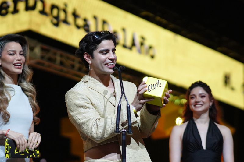 Aaron Mercury recibe el premio Líder Digital del Año en los Eliot Awards 2026 El creador de contenido Aaron Mercury en el escenario de los Eliot Awards 2026 sosteniendo con ambas manos la estatuilla dorada con forma de cubo que lleva la leyenda Eliot. Viste un saco corto en tono crema de cuello estilizado, mientras al fondo la conductora Galilea Montijo sonríe luciendo un vestido blanco de gala frente a las pantallas gigantes amarillas del evento.