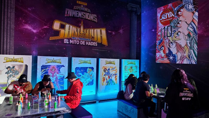 Asistentes reunidos en las mesas de actividades de la zona temática de Saint Seiya Next Dimension decorando a los Caballeros de Bronce con stickers y brillantina.