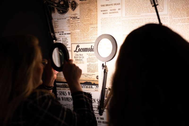 Visitantes utilizando lupas de aumento para leer titulares y notas impresas sobre portadas de periódicos históricos dedicados al hundimiento del Titanic.