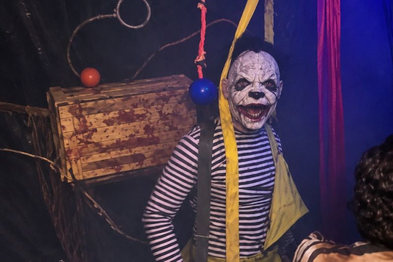 Caracterización en Freak Show Un actor caracterizado como un mico o payaso aterrador con máscara blanca desgastada, maquillaje siniestro y playera de rayas de pie en una escenografía de circo macabro.