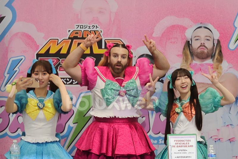 Integrantes del grupo Babybeard posando en el escenario frente a su lona promocional rosa; al centro, Ladybeard luciendo un traje rosa de magical girl con moños, flanqueado por las dos cantantes con trajes azules.
