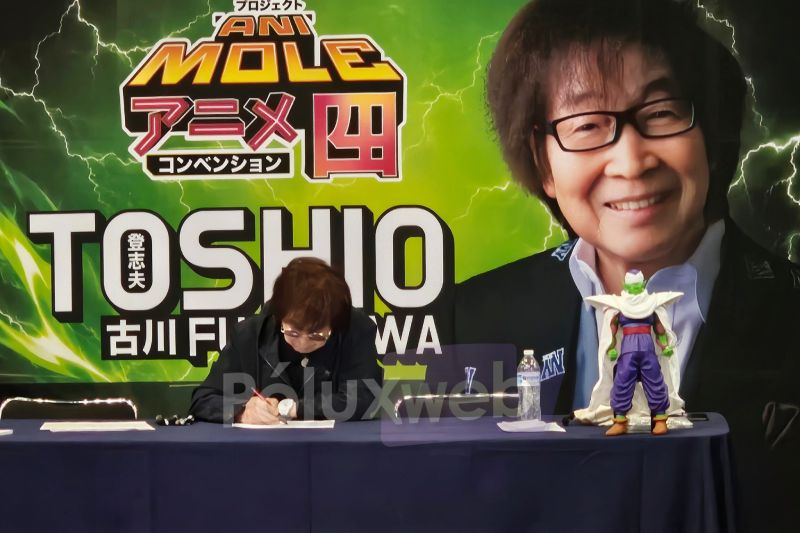 El actor de voz japonés Toshio Furukawa sentado en su mesa de autógrafos firmando documentos, frente a un banner con su rostro y con una figura de Piccolo colocada sobre la mesa.