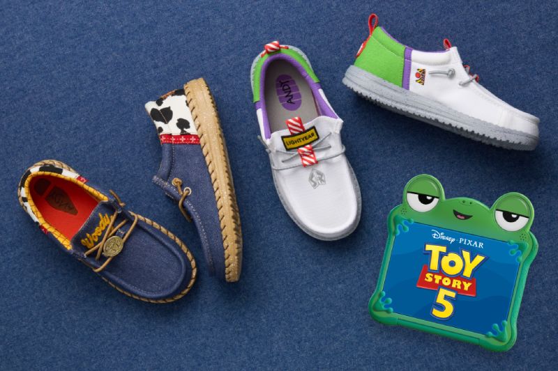 Colección de calzado casual HEYDUDE x Toy Story Fotografía de producto en vista superior cenital con cuatro modelos de calzado mocasín de la colaboración HEYDUDE x Toy Story, dispuestos sobre un fondo textil azul de mezclilla junto al logotipo oficial de la película en forma de juguete de rana verde.