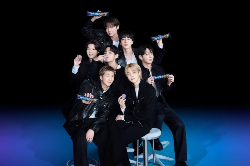 Fotografía promocional de los siete integrantes de BTS (RM, Jin, SUGA, j-hope, Jimin, V y Jungkook) vestidos con atuendos negros, posando juntos en un estudio con paquetes y galletas moradas de la edición limitada de OREO x BTS.