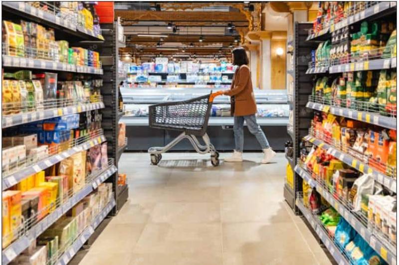 Pasillo de supermercado con consumidor en movimiento Fotografía de una persona empujando un carrito por un pasillo de supermercado con estantes llenos de productos empaquetados y una zona refrigerada al fondo.