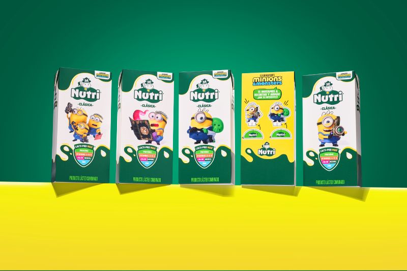 Fotografía de estudio que exhibe cinco empaques en formato tetra pack de la edición especial Nutri Clásica en colaboración con la película Minions & Monstruos. Cuatro envases presentan fondo blanco con verde y personajes como Minions sosteniendo cámaras o portando disfraces, mientras que el envase central muestra un diseño amarillo interactivo con instrucciones para recortar y armar figuras. La composición descansa sobre una superficie amarilla y un fondo verde.