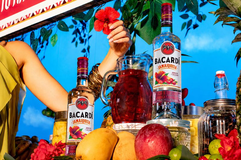 Botellas de cristal del ron saborizado Bacardí Jamaiquita expuestas en un puesto de frutas con flores rojas de jamaica, mangos y piñas. Al centro destaca una jarra de cristal con la bebida preparada de color rojo oscuro sobre un fondo de vegetación y cielo azul.