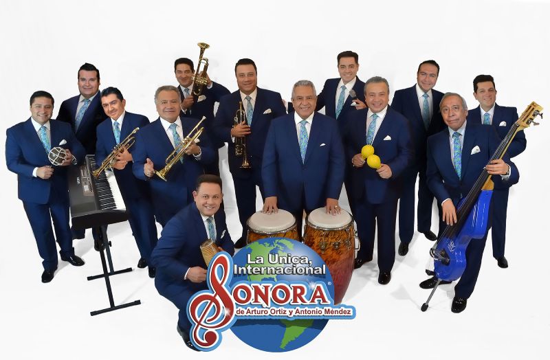 Fotografía promocional de una agrupación musical tropical con instrumentos y vestimenta formal.